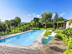 A swimming pool with a wooden deck, surrounded by lush greenery and a clear sky.