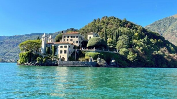 Un edificio grande de varios pisos con una cúpula destacada en el techo, ubicado en una isla junto al lago rodeada de abundante vegetación y colinas.