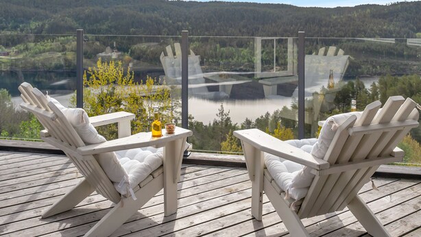 Two white Adirondack chairs on a wooden deck with a glass railing, overlooking a lake and forested hills. - Vacation home Solbu by Interhome (Eikerapen)