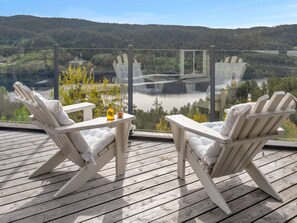 Two white Adirondack chairs on a wooden deck with a glass railing, overlooking a lake and forested hills. - Vacation home Solbu by Interhome (Eikerapen)