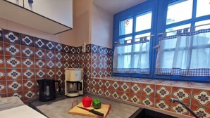 Una cocina con baldosas estampadas, una cafetera, una tabla de cortar con verduras y una ventana con cortinas.