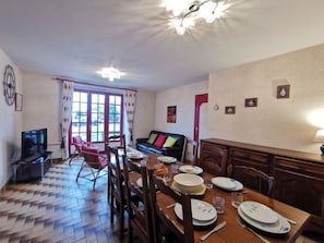 A dining area with a wooden table set for four, surrounded by chairs. There is a television on the left and a sofa in the background.