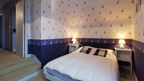 Un dormitorio con cama, dos mesitas de noche y una mesita auxiliar con lámparas. Las paredes tienen un diseño con barcos a vela.