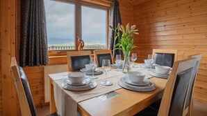 Et spisekammer i en træhytte med et bord dækket til fire personer, store vinduer og udsigt over landskabet.
