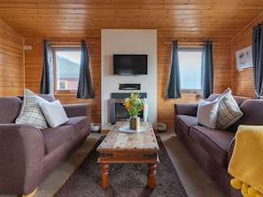 A cozy cabin interior with two sofas facing a central wooden coffee table, a flat-screen TV mounted on the wall, and a window with curtains.