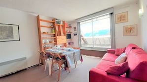 A dining area with a table set for four, wooden chairs, a pink sofa, and a large window with a view of a balcony. - Cozy Studio for 6 with Mountain View, Ski Locker, and Modern Amenities in Piau-Engaly (Piau-Engaly)