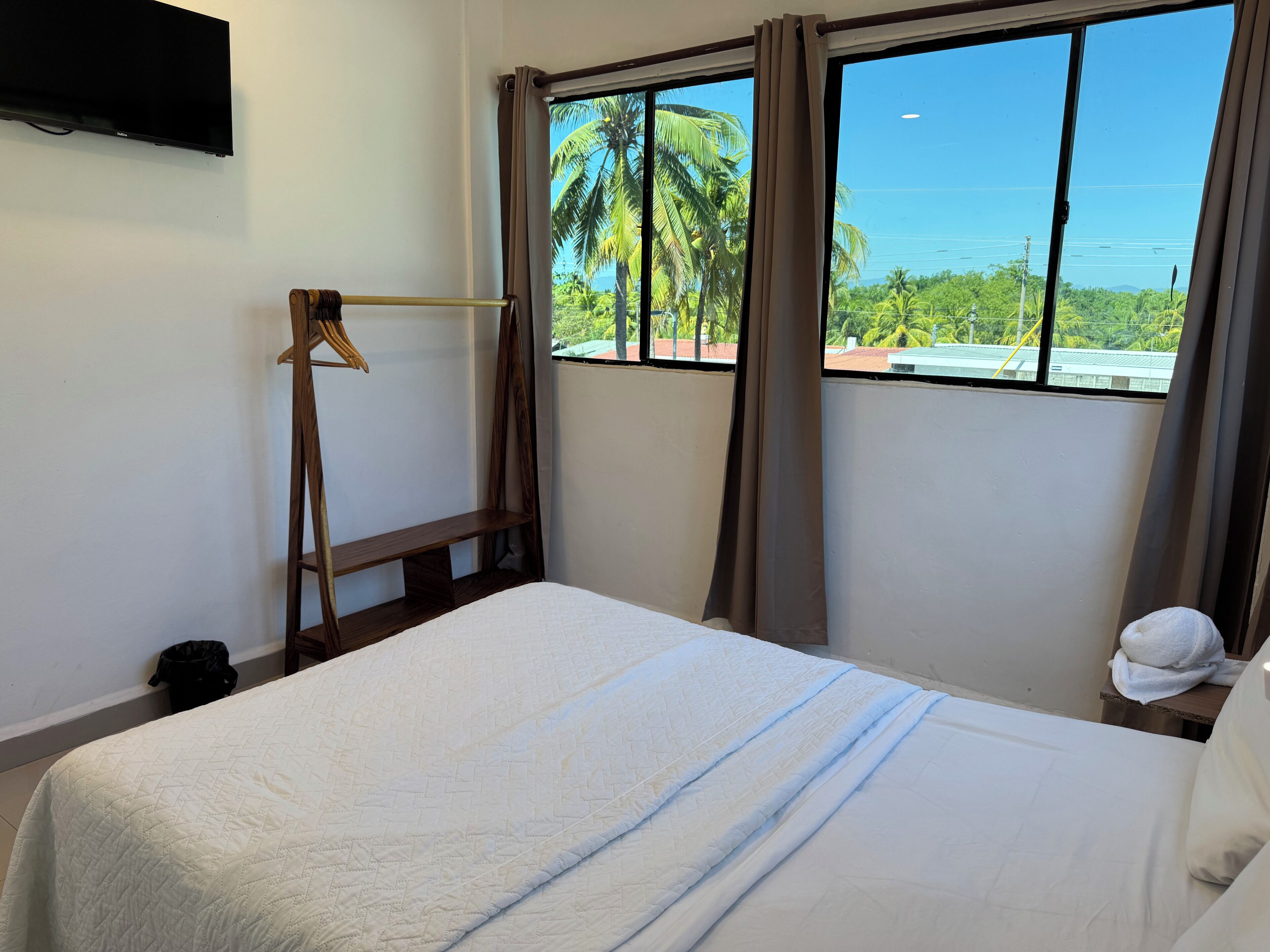 Ein Hotelzimmer mit Bett, einem an der Wand befestigten Fernseher, einem Fenster mit Vorhängen und Blick auf Palmen und das Meer.