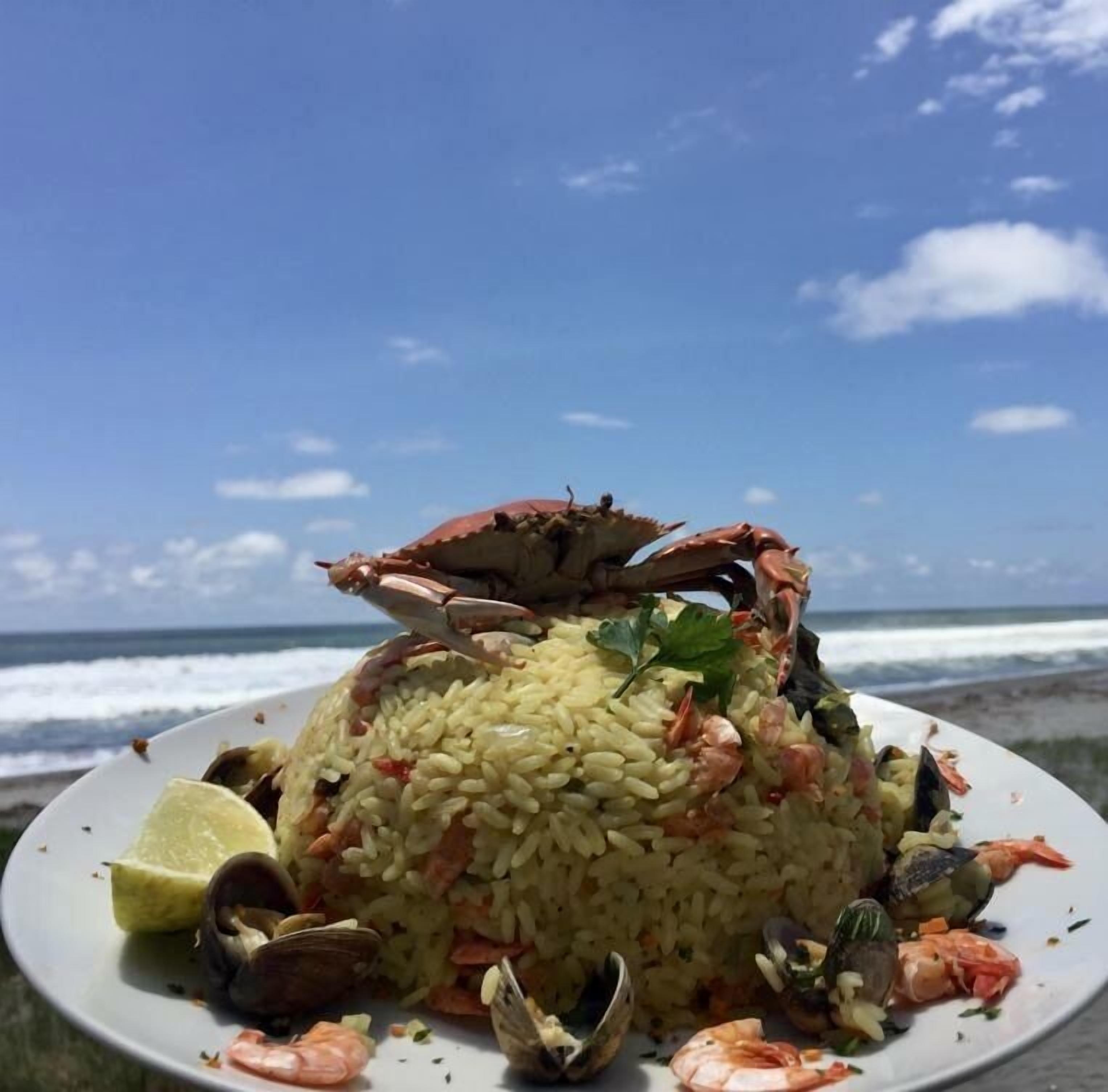 A seafood rice dish on a plate with clams and shrimp, set against a beach backdrop.
