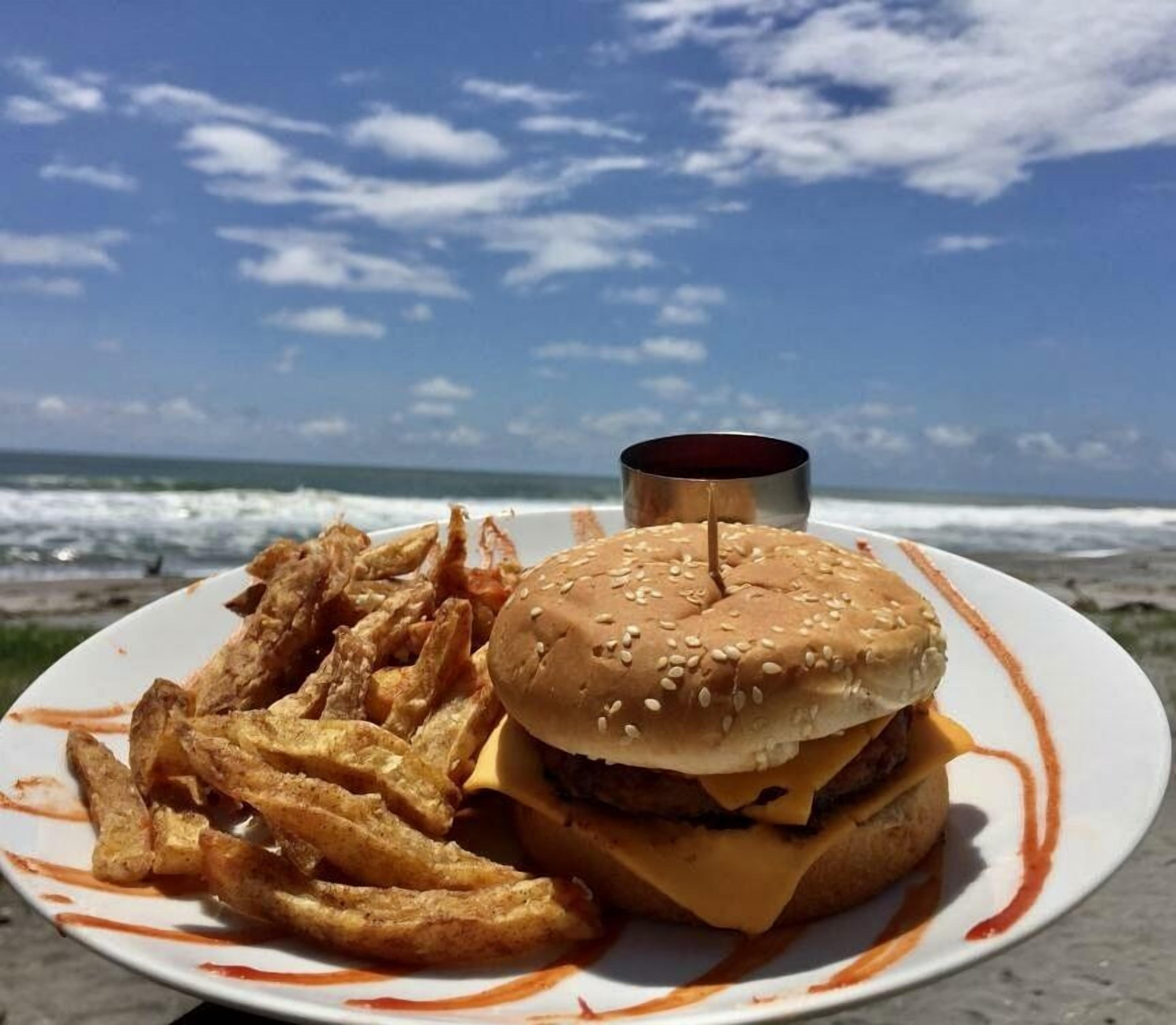 En tallrik med en hamburgare och pommes frites på en strand, med havet i bakgrunden.