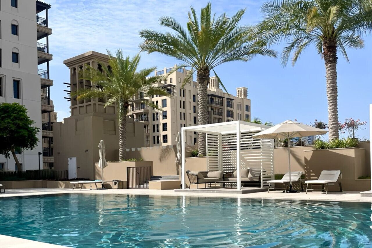 A swimming pool with lounge chairs and umbrellas, surrounded by palm trees and modern buildings.