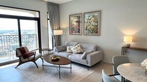 Een moderne woonkamer met een grijze bank, een ronde salontafel, een fauteuil en een eettafel met een lamp. Twee ingelijste kunstwerken hangen aan de muur en er is een groot raam met uitzicht.