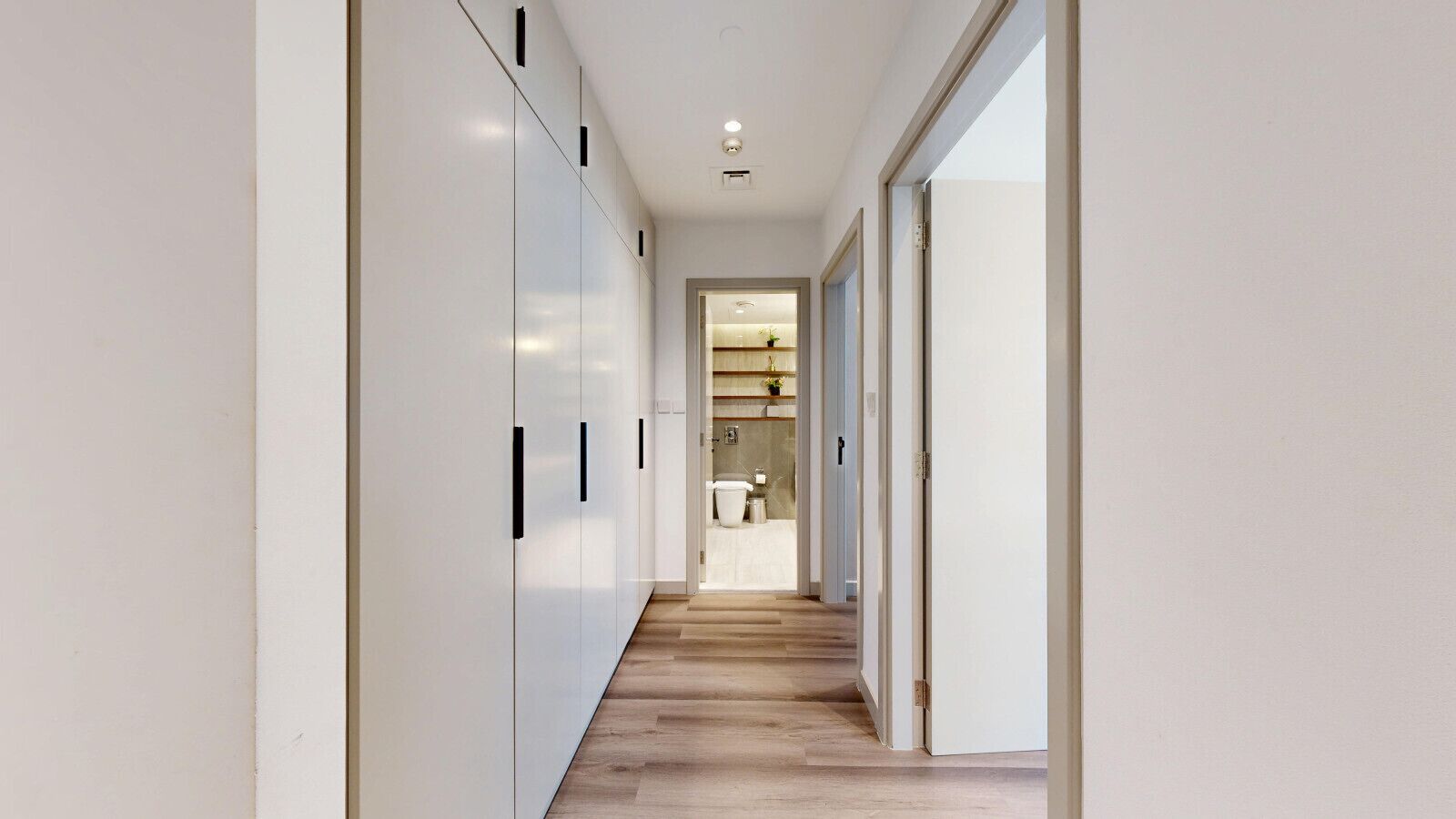 A modern hallway with white walls, wooden flooring, and a view into a bathroom with a toilet and shelves.