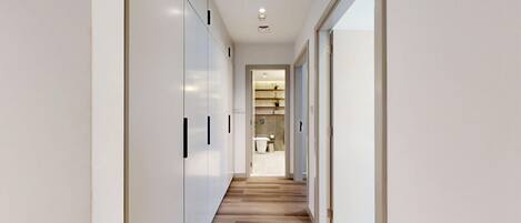 A modern hallway with white walls, wooden flooring, and a view into a bathroom with a toilet and shelves.