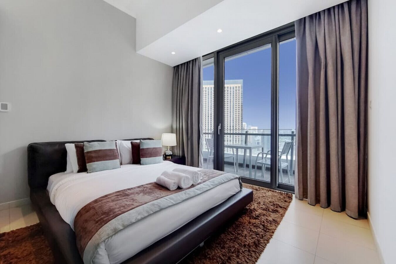 A modern hotel room with a large bed, a view of the city, and a balcony with a table and chairs.
