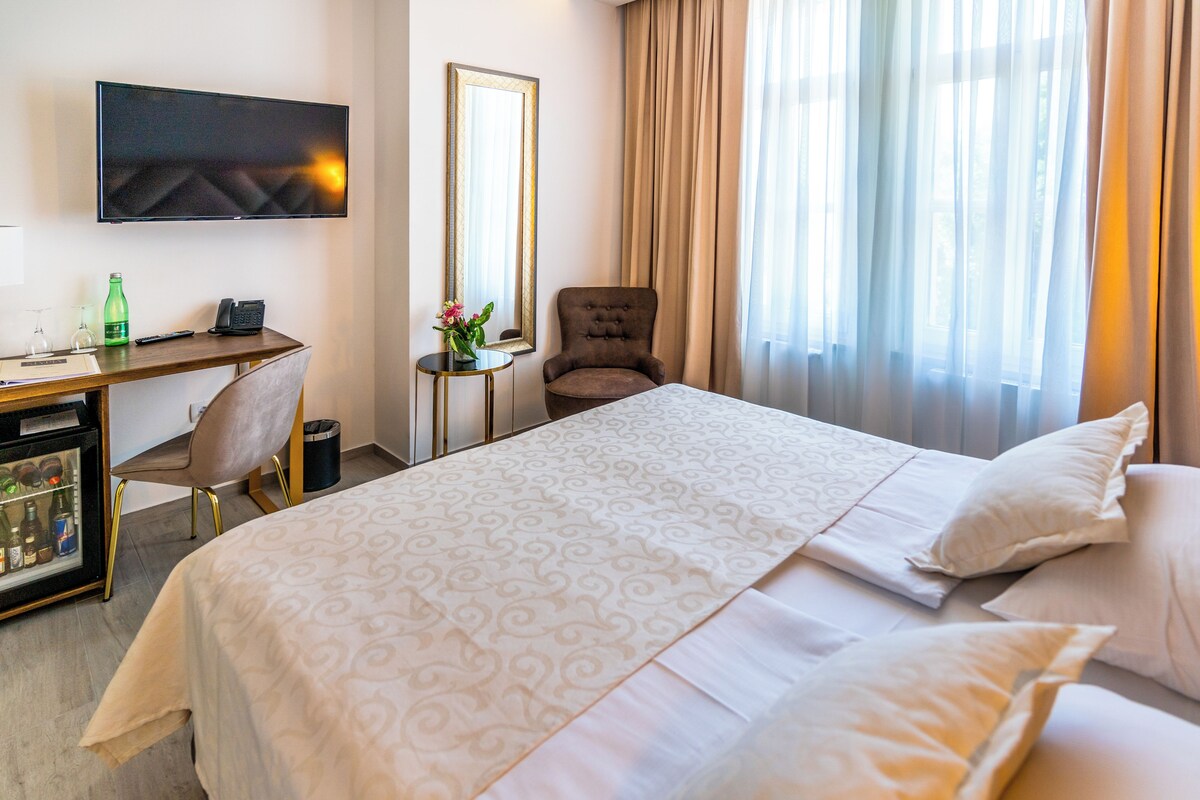 Habitación de hotel con cama, escritorio con computadora, una silla, televisor montado en la pared y ventana con cortinas.