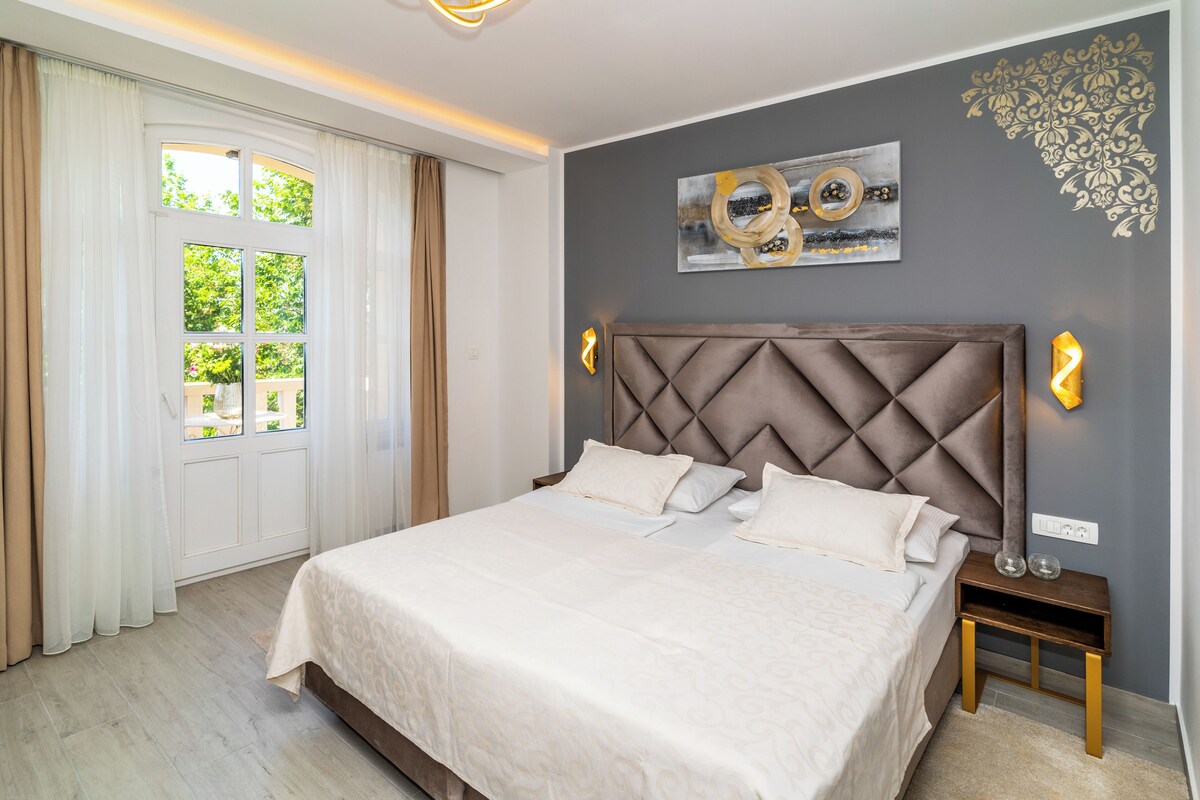 Une chambre à coucher avec un grand lit, une tête de lit, une table de chevet, une lampe fixée au mur et une fenêtre avec des rideaux.