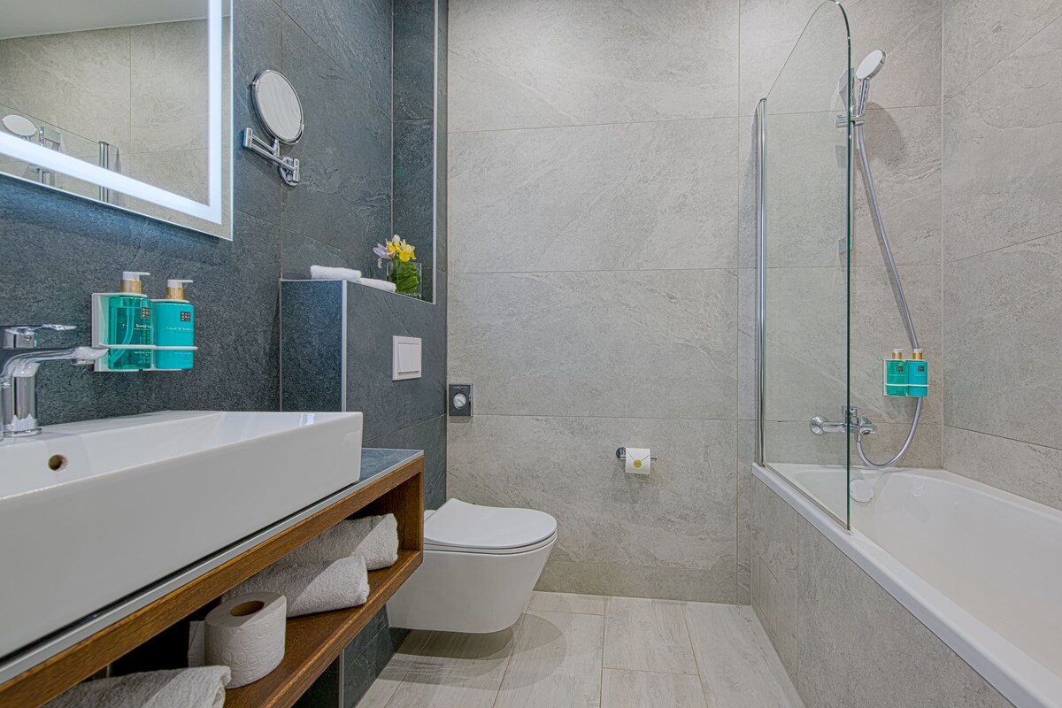 Une salle de bain moderne avec un lavabo blanc, un miroir et des toilettes, une douche à parois de verre et une étagère en bois où sont posées des serviettes.