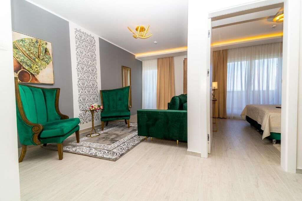 A hotel room with a green armchair, a green sofa, a small side table with flowers, a bed with a beige bedspread, and a large window with curtains.