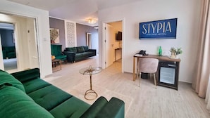 Une chambre d’hôtel moderne avec un canapé vert, une petite table ronde, un bureau avec une chaise et un grand écran de télévision affichant le nom de l’hôtel.