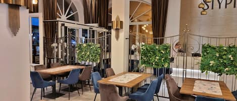 Un restaurante con mesas de madera y sillas azules, rodeado de barandillas metálicas decorativas y follaje verde.