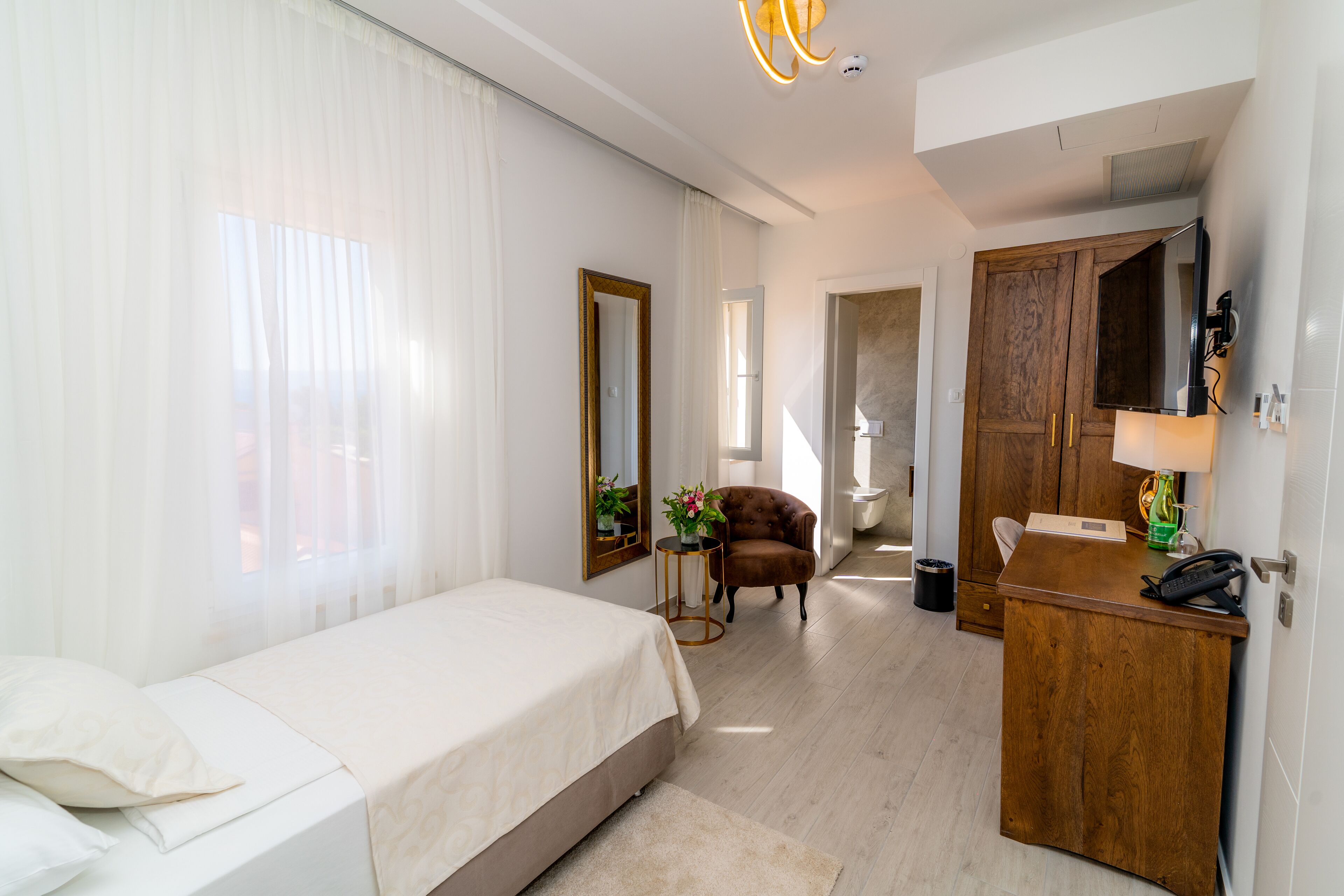 A hotel room with a bed, a wooden desk, a chair, a mirror, and a small table with a lamp.