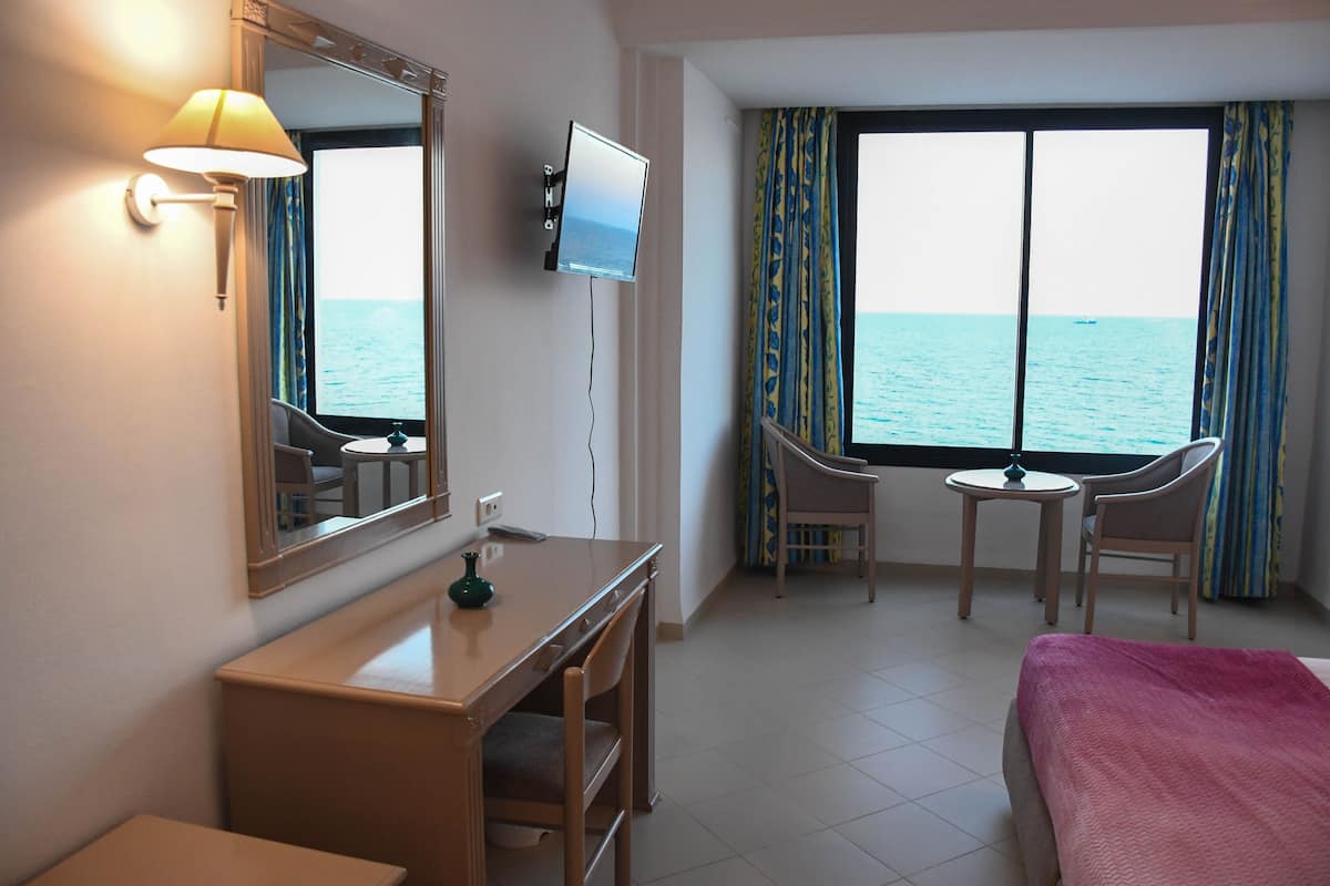 A hotel room with a bed, a desk, a chair, a TV, and a window with a view of the sea.