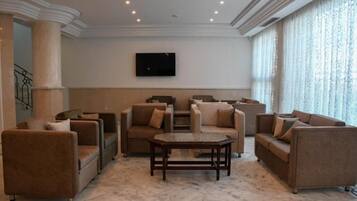 Amplia sala de estar con varios sofás y una mesa de centro, un televisor de pantalla plana montado en la pared y ventanas grandes con cortinas translúcidas.