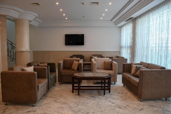 A spacious living area with multiple sofas and a coffee table, a flat-screen TV mounted on the wall, and large windows with sheer curtains. - Hôtel Maravilla Sousse (Sousse)