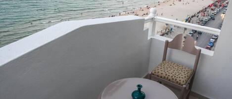 A balcony with a view of a beach and a pier. There is a chair and a small table with a vase on it.