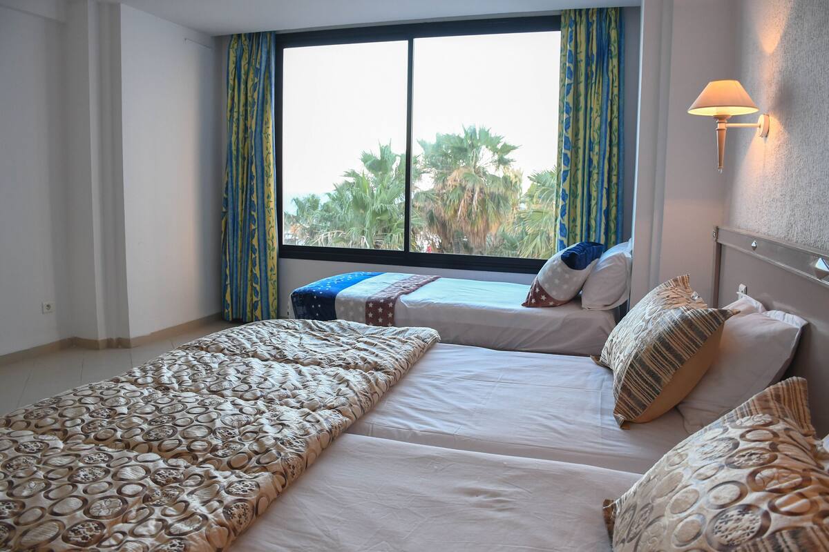 A hotel room with two beds, a large window with curtains, a wall-mounted lamp, and a view of palm trees outside.