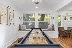 Una mesa de shuffleboard en una habitación con ventanas amplias, una chimenea y una zona de estar con cojines.