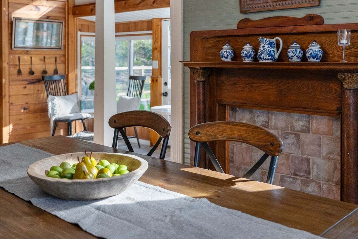 A wooden dining table with a bowl of fruit, a fireplace with blue and white ceramic pots, and a wooden chair.