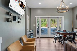 Una sala de estar moderna con televisor de pantalla plana, consolas de videojuegos, un sofá, una mesa de comedor con sillas y una araña.