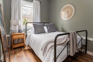 Un dormitorio con cama, mesita de noche, lámpara y ventana con cortinas.