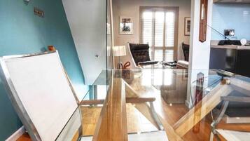 A modern meeting room with a glass table, wooden flooring, a whiteboard, and a window with blinds.