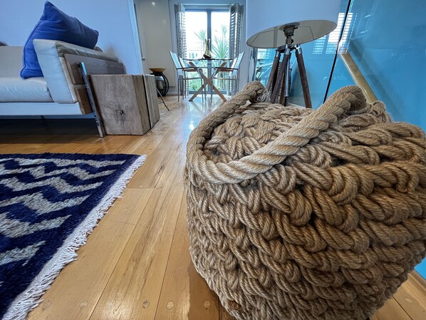 A living room with a large beige rope-woven pouf, a blue and white rug, a white sofa, and a dining area with a table and chairs.