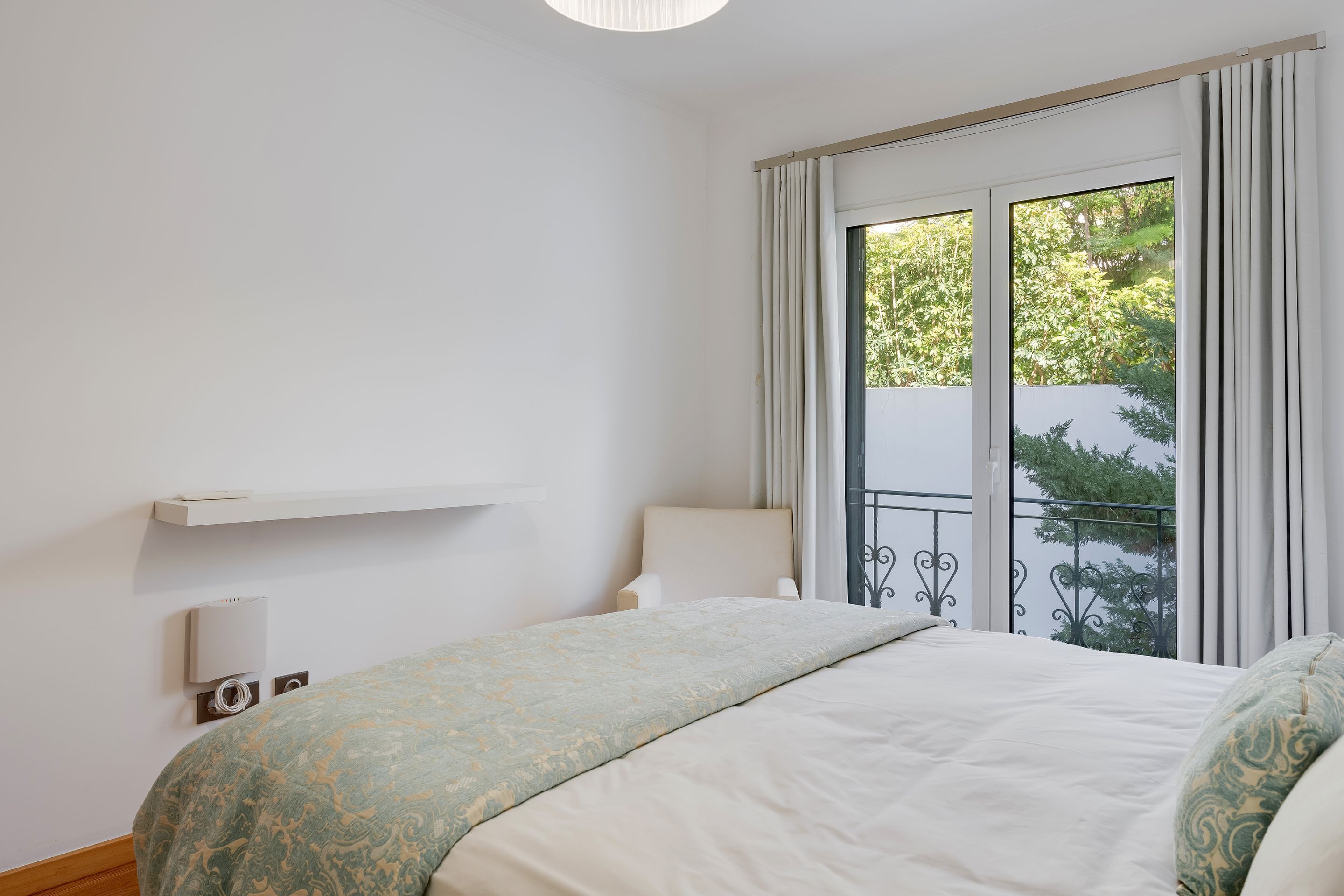 Un dormitorio con cama, una silla, un ventanal con cortinas y un balcón.