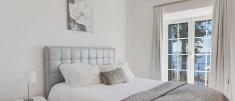 Un dormitorio con cama, mesita de noche, lámpara, ventana con cortinas y vista al mar.