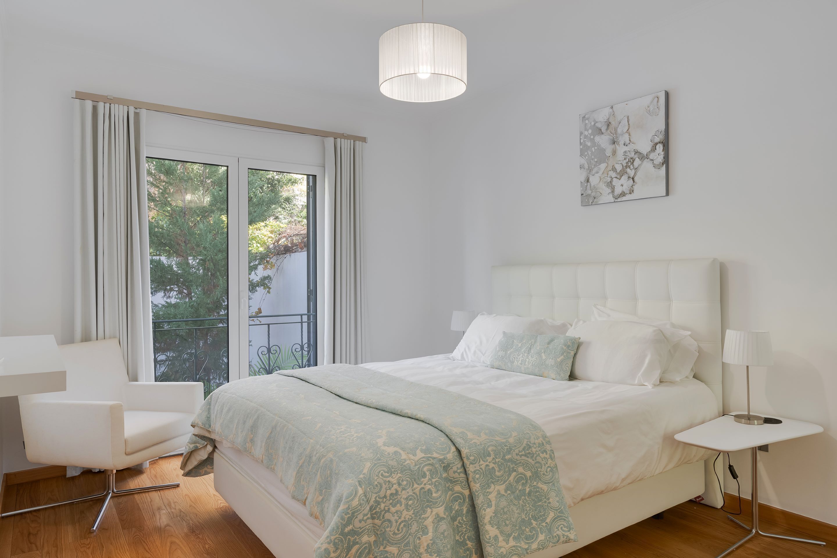 A modern bedroom with a large bed, a chair, a bedside table, and a view of greenery outside.