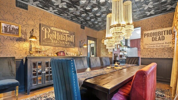 A dining area with a wooden table, upholstered chairs, a chandelier, and a wall with a sign. - The Prohibition -Uniquely Designed Midtown Stunner (Kansas City)