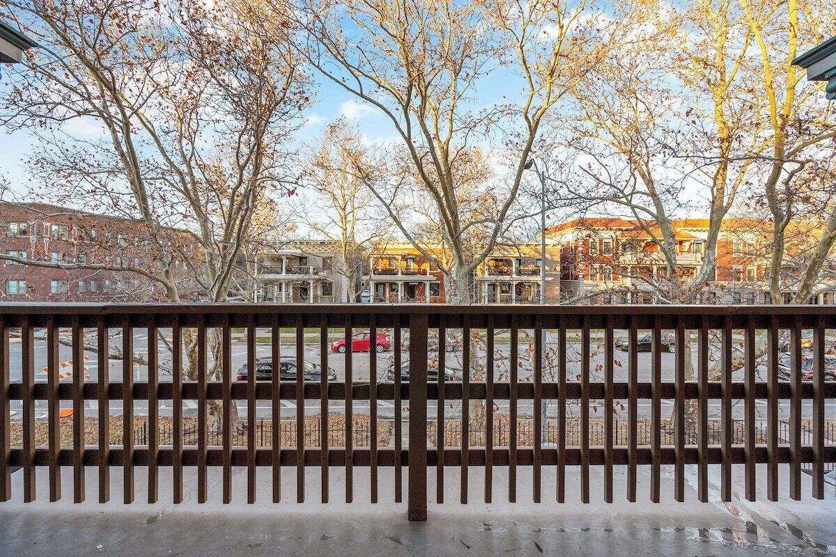 A view from a balcony with a metal railing, overlooking a parking lot and buildings with brick facades.