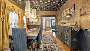 A dining area with a wooden table, chairs, and a chandelier. There is a bar area with a counter and stools. The room has patterned wallpaper and a decorative ceiling. - The Prohibition -Uniquely Designed Midtown Stunner (Kansas City)