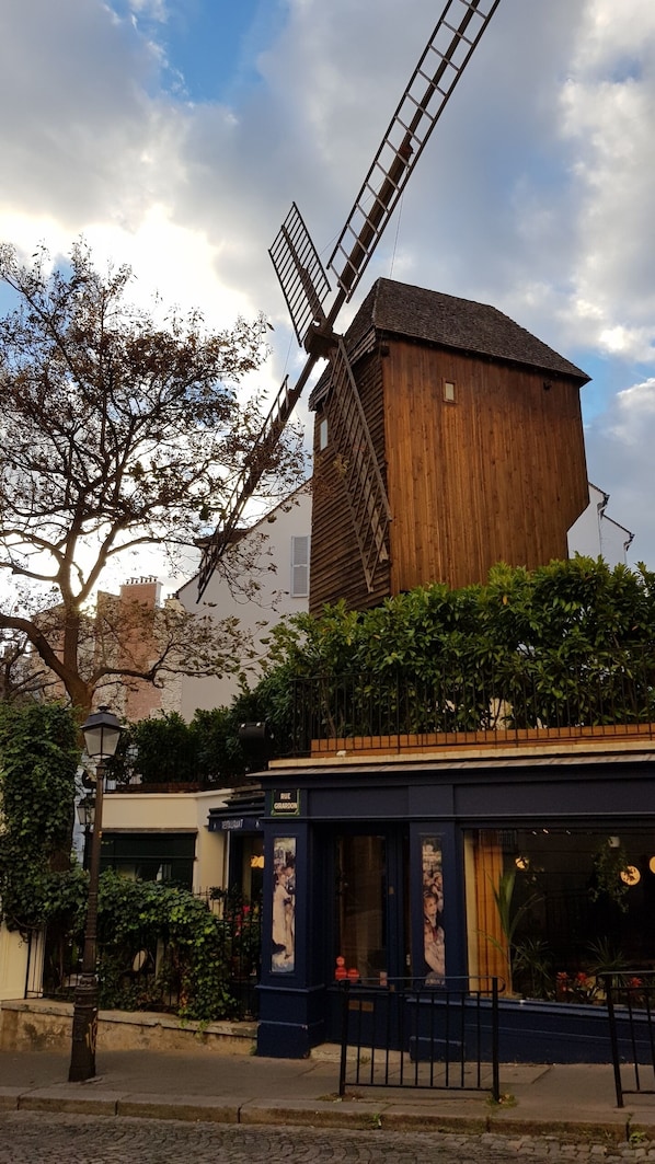 A traditional windmill with a wooden structure and a slanted roof, adjacent to a building with a dark facade and a sign reading "RUE GIRARDON".