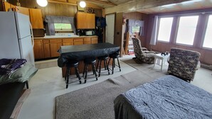 Un interior de cabaña acogedor con techo de madera, una zona de cocina con armarios de madera, una mesa de comedor con taburetes, un sillón estampado y vistas a través de amplias ventanas.