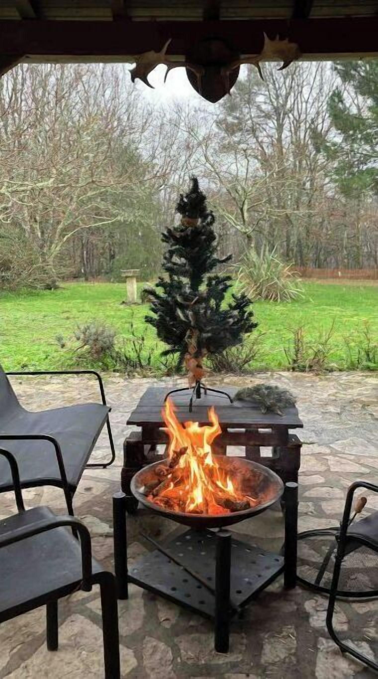 A fire pit with burning logs, surrounded by outdoor chairs, set on a stone patio with a view of a forested area.