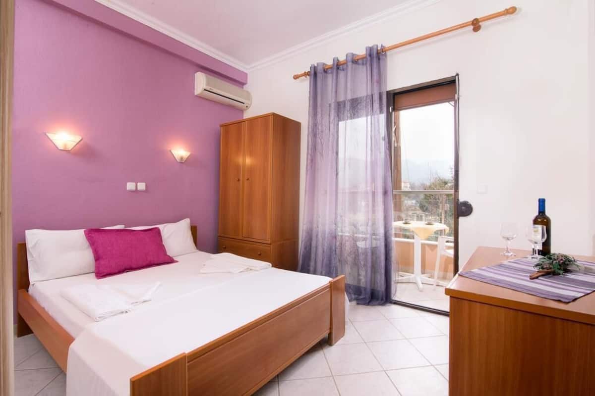 A bedroom with a bed, a wooden wardrobe, a balcony with a table and chairs, and a view of the outdoors.