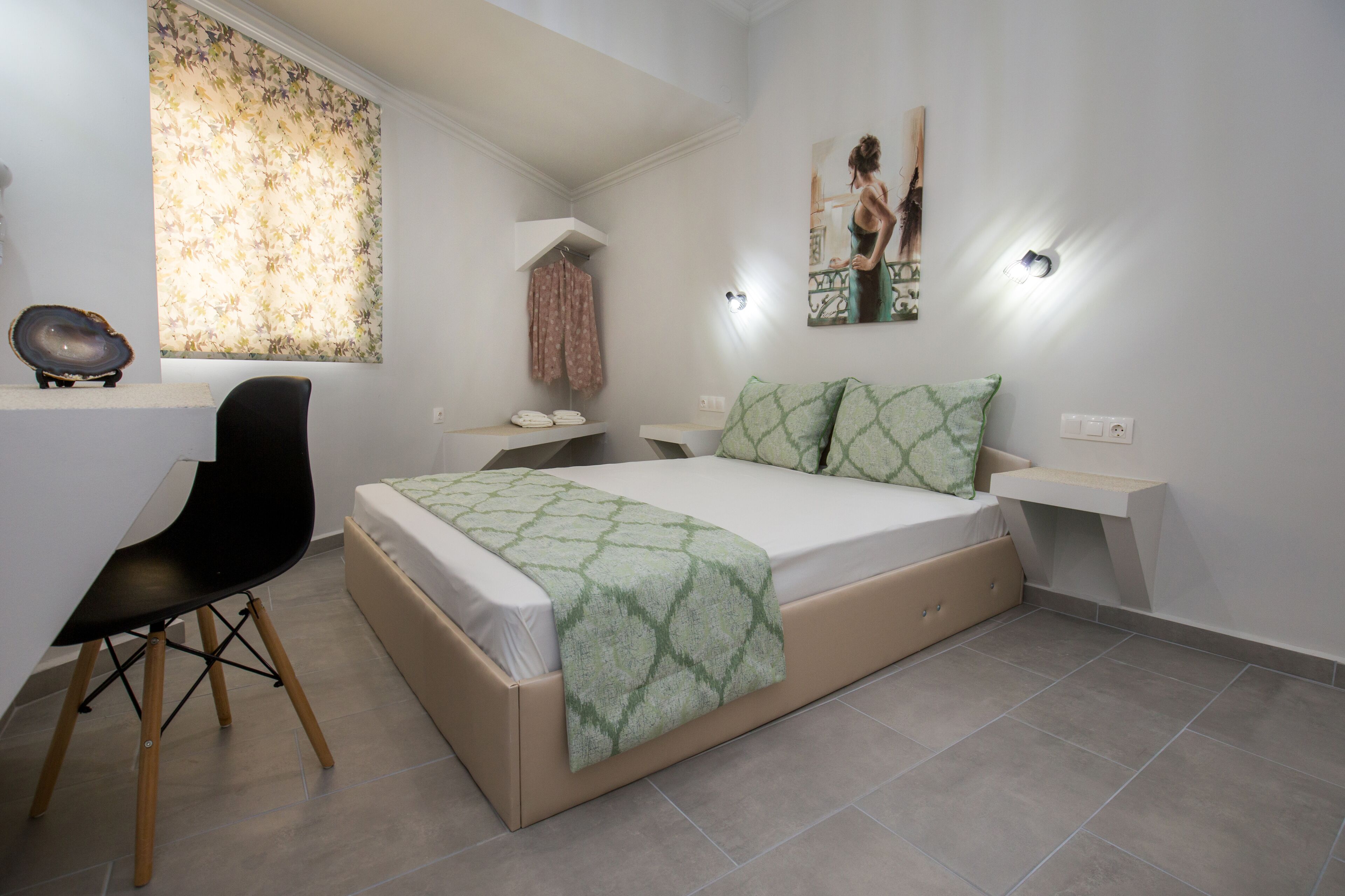 A bedroom with a bed, a desk, a chair, a window with curtains, a wall-mounted light, and a painting.