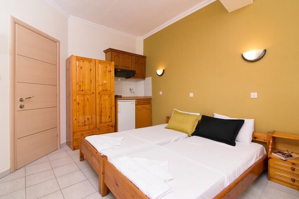 A bedroom with a wooden bed, a nightstand, a wardrobe, and a kitchen area with wooden cabinets and a stove.