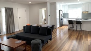 A modern living room with a dark sofa, a wooden coffee table, and a grey ottoman. A kitchen with white cabinets and a stainless steel refrigerator is visible through an open door. - House in Belmont fully renovated (Belmont)