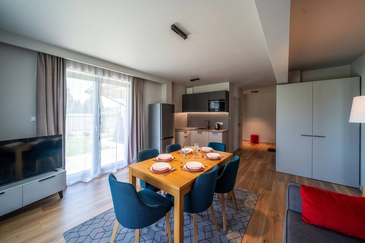 Uma sala de estar moderna com uma mesa de jantar posta para seis pessoas, uma televisão e um sofá com um travesseiro vermelho.